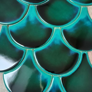 May include: Close-up of teal-colored, fan-shaped tiles arranged in a pattern. The tiles have a glossy finish and a gradient effect, transitioning from dark teal to lighter shades. The tiles are likely for decorative use.
