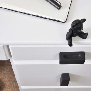 Op de afbeelding: Een witte bureau met zwarte lade grepen en een zwart teddybeer beeldje. Een zwarte pen en een notitieboek liggen op het bureau. Het bureau heeft twee laden met rechthoekige zwarte handgrepen.