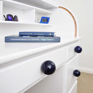 May include: White desk with a roll-top design, featuring dark blue spherical drawer pulls. The desk has shelves with books, sunglasses, and a blue digital clock. A blue and white vase and a plant are on top.