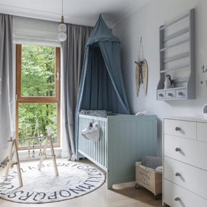 Pu&ograve; includere: Una cameretta per bambini con una culla azzurra, un com&ograve; bianco e un tappeto rotondo con lettere dell'alfabeto. Una zanzariera &egrave; appesa sopra la culla. La stanza ha una finestra con tende grigie e un telaio in legno. Le parole "Let's fly to the moon" sono sul muro.