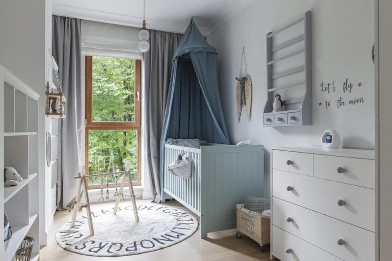 Pu&ograve; includere: Una cameretta per bambini con una culla azzurra, un com&ograve; bianco e un tappeto rotondo con lettere dell'alfabeto. Una zanzariera &egrave; appesa sopra la culla. La stanza ha una finestra con tende grigie e un telaio in legno. Le parole "Let's fly to the moon" sono sul muro.