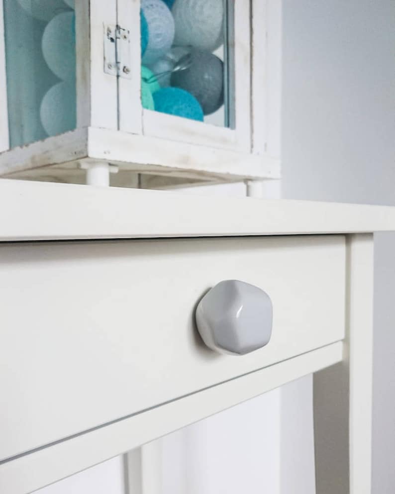 Puede incluir: Mesita de noche blanca con un solo caj&oacute;n y una perilla de cer&aacute;mica blanca. El caj&oacute;n est&aacute; cerrado.