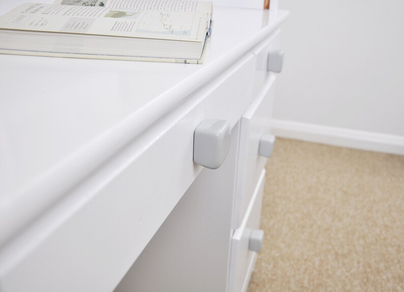 Peut inclure: Une commode blanche avec quatre tiroirs et des poign&eacute;es de tiroir en plastique gris. La commode est en bois et a une finition blanche lisse.