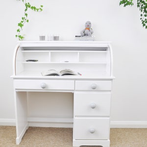 May include: White roll top desk with three drawers and a shelf. The desk has a light wood top and white knobs on the drawers. The desk is sitting on a beige carpet.