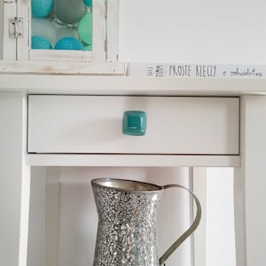 Puede incluir: Mesita de noche blanca con un cajón y un tirador de cajón de color turquesa. La mesita de noche tiene un estante debajo del cajón. Una jarra de espejo plateado se encuentra en el estante. La mesita de noche tiene un pequeño letrero en la parte superior que dice "Just.  Timm.  Proste Rzeczy.  o rodzicielstwie"