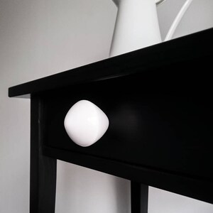 Puede incluir: Una mesa auxiliar de madera negra con un pomo de cerámica blanca en el cajón. Una jarra blanca se encuentra encima de la mesa.