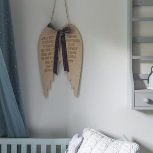 Pu&ograve; includere: Decorazione murale ad ali d'angelo in legno con nastro scuro e testo. Le ali sono appese a una parete bianca. &Egrave; visibile una mensola grigia con una statuina di pinguino e cassetti. Una culla blu e una tenda trasparente sono sullo sfondo.