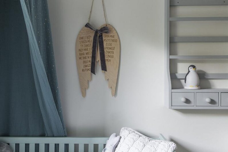 Pu&ograve; includere: Decorazione murale ad ali d'angelo in legno con nastro scuro e testo. Le ali sono appese a una parete bianca. &Egrave; visibile una mensola grigia con una statuina di pinguino e cassetti. Una culla blu e una tenda trasparente sono sullo sfondo.