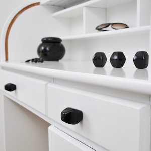 Könnte beinhalten: Ein weißer Schreibtisch mit Roll-Top-Design, mit schwarzen geometrischen Schubladengriffen und dekorativen Gegenständen. Der Schreibtisch hat ein Regal mit Sonnenbrillen und drei schwarzen geometrischen Objekten. Ein schwarzer Topf und eine Perlenkette befinden sich auf der Schreibtischoberfläche.