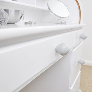 May include: White vanity desk with four drawers and ceramic knobs. The desk has a white top with a shelf above the drawers. A small round mirror is on the top shelf.
