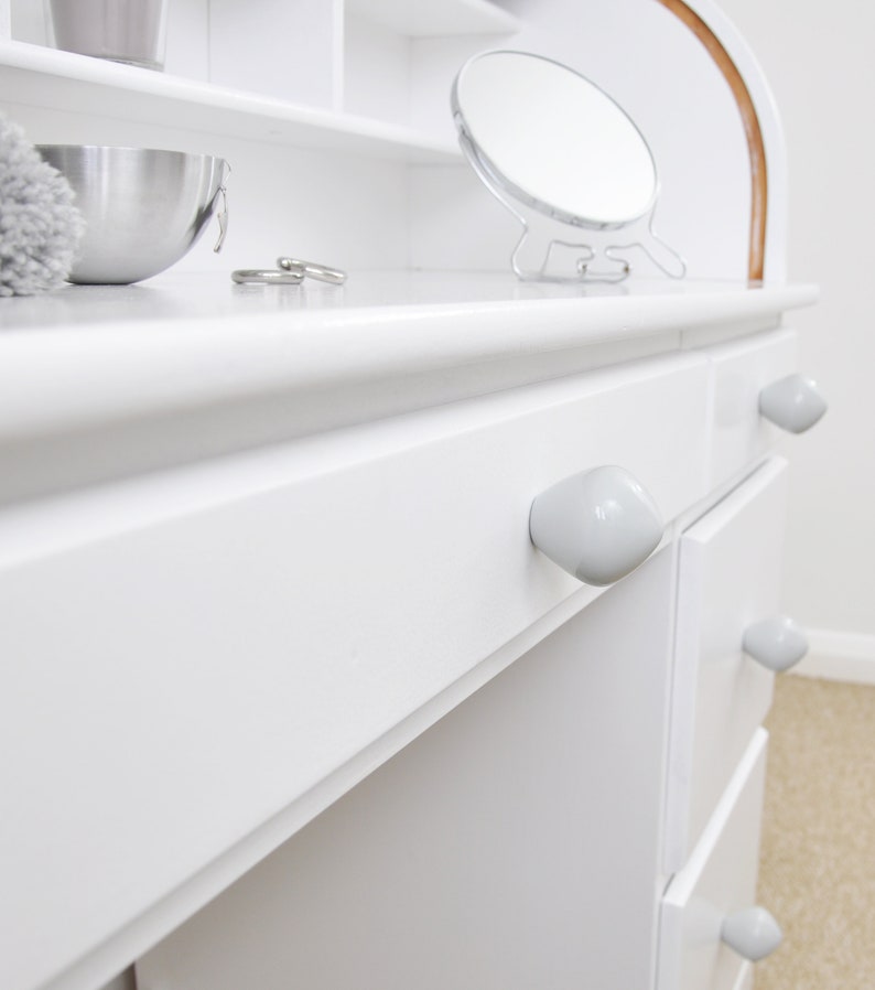 May include: White vanity desk with four drawers and ceramic knobs. The desk has a white top with a shelf above the drawers. A small round mirror is on the top shelf.