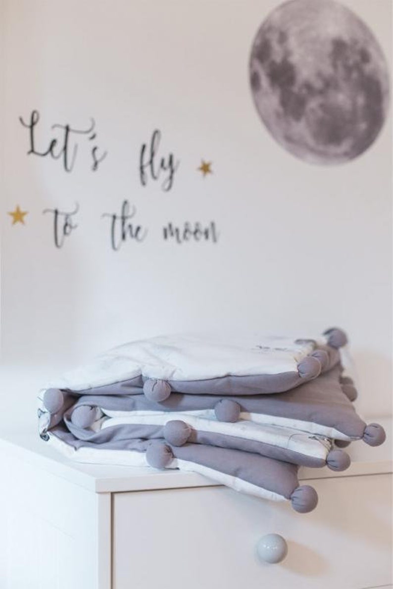 Pu&ograve; includere: Una coperta per bambini grigia e bianca piegata con pompon grigi sui bordi. La coperta &egrave; impilata su un com&ograve; bianco. Il muro dietro la coperta ha un adesivo a forma di luna e la scritta "Let's fly to the moon".