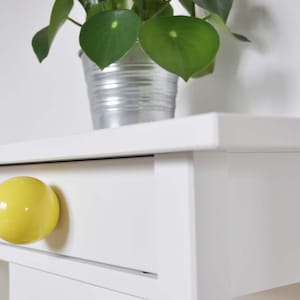 May include: A white bedside table with a yellow round knob on the drawer. A potted plant sits on top of the bedside table.