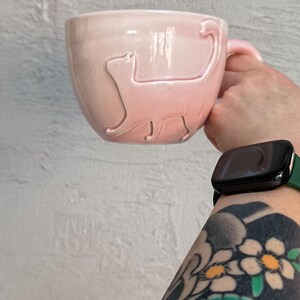Op de afbeelding: Een roze keramische mok met een katten silhouet ontwerp. De mok heeft een handvat en wordt tegen een witte, getextureerde muur gehouden. De mok is lichtroze.