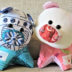Pode incluir: Dois brinquedos de pelúcia feitos à mão com a forma de porcos. O porco à esquerda é azul e branco com um padrão floral e o porco à direita é rosa e branco com um padrão floral. Ambos os porcos têm olhos pretos e um nariz vermelho.