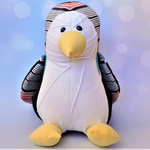 Pode incluir: Um brinquedo de pelúcia de pinguim com listras pretas e brancas, detalhes em rosa e azul e um bico e pés amarelos.