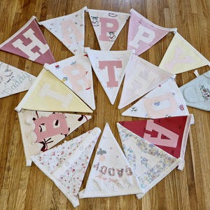 May include: A circular arrangement of fabric pennant flags. The flags display letters, patterns, and the words "Daddy" and "Love you". The flags are in various colors, including pink, yellow, and red. The flags are made of fabric.
