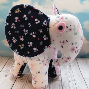 May include: A stuffed elephant toy with a floral pattern. The elephant's ears are navy blue with small flowers, while the body is white with pink and blue flowers. It has a pink eye and black pupil.