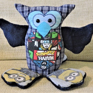 May include: A blue and black owl plush toy with a blue beak and black eyes. The owl is wearing a plaid shirt with a Marvel Comics logo on the front. The owl has two feet that are shaped like Minions.