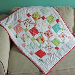 May include: A white baby quilt with a red, green, and white patchwork design. The quilt is made of squares of different fabrics and is edged with a red trim.