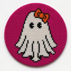 May include: A circular cross-stitch embroidery piece featuring a white ghost with black eyes and a bright orange bow on its head, set against a vibrant magenta background. The ghost has a scalloped bottom edge and is outlined in black thread.