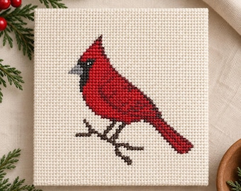 May include: A square piece of off-white Aida fabric featuring a cross-stitch design of a bright red cardinal bird with a black face mask and a gray beak, perched on a dark brown branch, rendered in a grid-like pattern of individual stitches.