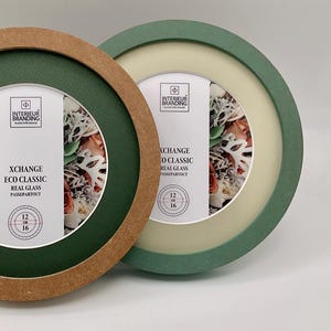 May include: Two round picture frames, one with a brown border and the other with a green border. The frames have a white mat and a floral image inside. Text on the frame reads "XCHANGE ECO CLASSIC REAL GLASS PASSEPARTOUT" and "12 cm 16 cm".