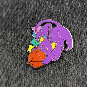 May include: A purple panther enamel pin with yellow accents, clutching an orange and black polyhedral die. The pin features a geometric design with colorful shapes in the background. The pin has a silver outline.
