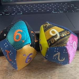 May include: Two multi-sided fabric dice featuring various textures and colors including teal, yellow, blue, black, and pink. Each triangular panel is stitched together and displays embroidered numbers in contrasting thread colors like orange and white.