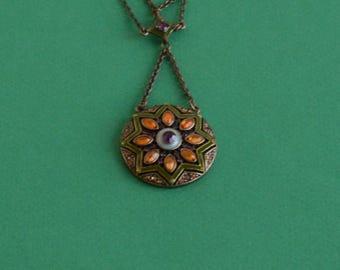 May include: A bronze-toned metal pendant necklace featuring a circular star-shaped design. The center contains a small purple stone surrounded by a ring of orange marquise-shaped accents and green enamel detailing, suspended from a delicate chain.