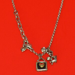 May include: Silver necklace with a charm in the shape of a small square with a heart inside. The heart is gold with a black background. The necklace also has a small silver heart charm and a silver crown charm with a clear crystal.