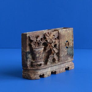 May include: A rectangular brown stone vase featuring intricate relief carvings of a potted flower and foliage on one side. The object has a flat base with cut-out details and a small square opening on the right side, showcasing a mottled texture.