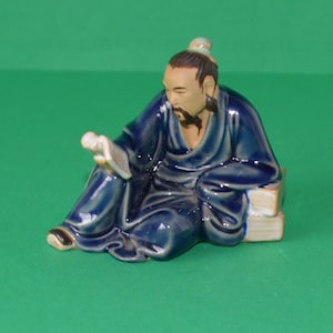 May include: A ceramic figurine of a seated Asian scholar in a blue robe, reading a scroll. The figure has a black beard and is seated on a stack of books. The figurine is on a green background.