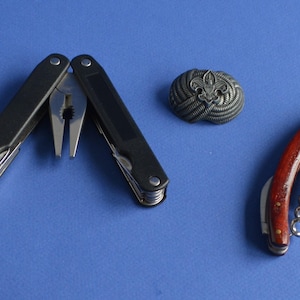 May include: A black multi-tool with pliers in an open position, a silver-toned metal emblem featuring a fleur-de-lis design, and a folding corkscrew tool with a reddish-brown wooden handle and silver metal components arranged on a blue surface.