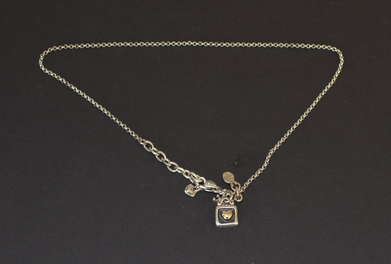 May include: A silver chain necklace with a small square pendant featuring a gold heart. The pendant has a black background and is attached to the chain with a silver clasp.