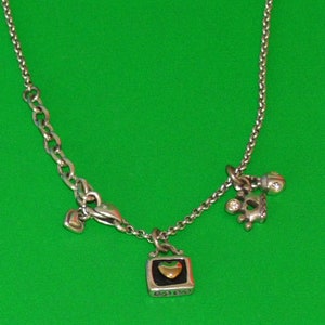 May include: A silver chain necklace with three charms: a small heart, a crown with a clear stone, and a square charm with a gold heart inside.