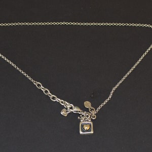 May include: A silver chain necklace with a small square pendant featuring a gold heart. The pendant is attached to a silver charm with a heart shape.