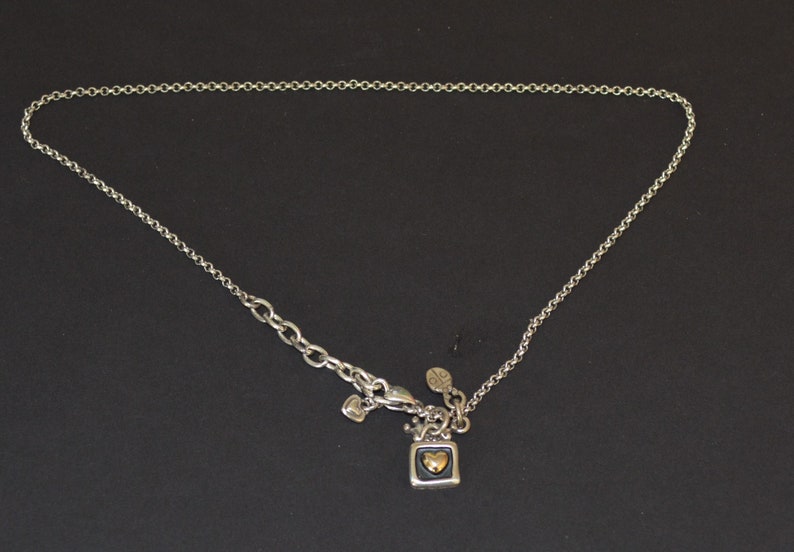 May include: A silver chain necklace with a small square pendant featuring a gold heart. The pendant is attached to a silver charm with a heart shape.