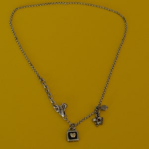 May include: A silver chain necklace with a silver charm in the shape of a padlock with a gold heart inside. The necklace also has two smaller charms, one shaped like a crown and the other like a ladybug.