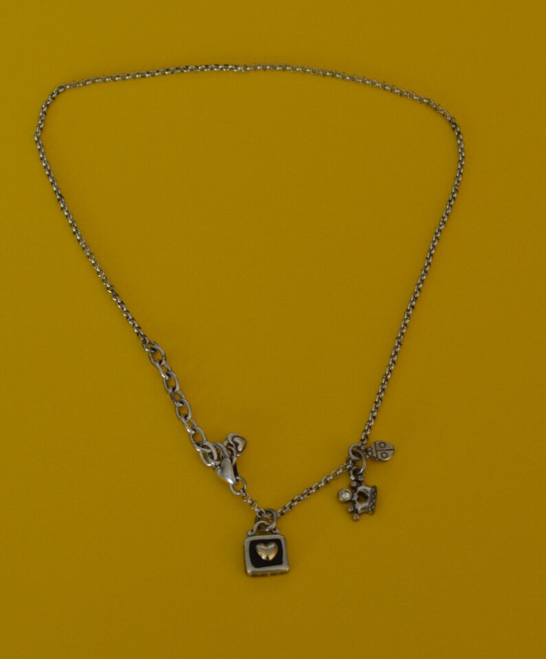 May include: A silver chain necklace with a silver charm in the shape of a padlock with a gold heart inside. The necklace also has two smaller charms, one shaped like a crown and the other like a ladybug.