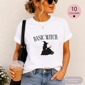 May include: A white short-sleeve t-shirt featuring the black text BASIC WITCH above a black silhouette graphic of a witch holding a drink next to a pumpkin. The shirt has a crew neckline and is worn with blue denim jeans and a black shoulder bag.