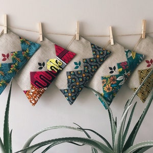 Peut inclure: Une rangée de pochettes en tissu décoratives avec des motifs géométriques et floraux colorés est suspendue à une ficelle avec des pinces à linge en bois. Les pochettes sont en lin naturel et sont accrochées contre un mur blanc, avec des plantes d'aloe vera au premier plan.