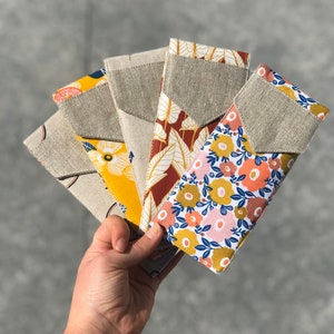 Op de afbeelding: Een hand houdt een waaier van vijf stoffen etuis vast. Elk etui heeft een linnen zak en een patroonklep. De patronen omvatten gele bloemen, rode bladeren en roze bloemmotieven. De etuis zijn ontworpen om brillen of andere kleine items te bevatten.