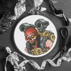 Puede incluir: Un bastidor de bordado de punto de cruz con una imagen pixelada de una calavera roja con ojos verdes y un soldado futurista. La obra de arte está sobre un fondo blanco, rodeada de materiales de artesanía como tijeras y cinta métrica.