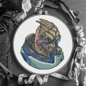 Puede incluir: Un retrato de punto de cruz de un personaje del videojuego Mass Effect, enmarcado en un bastidor blanco. El personaje tiene un diseño detallado y pixelado con tonos de azul, beige y morado. El fondo incluye suministros de costura y elementos decorativos.