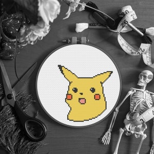 Könnte beinhalten: Ein Kreuzstichrahmen mit einem gelben Pikachu-Design mit orangefarbenen Wangen und offenem Mund. Der Stickrahmen ist von Bastelzubehör umgeben, darunter eine Schere, Faden und ein Maßband.