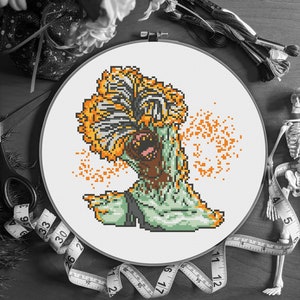 May include: A completed cross-stitch project featuring a pixelated monster with an orange frill and green body. The design is set in a white hoop, with orange specks around the monster. Measuring tape and embroidery floss are visible.