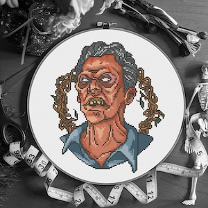 Op de afbeelding: Een kruissteekportret van een zombie-achtig figuur met grijs haar, een bebloed gezicht en een blauw overhemd, geplaatst in een witte hoepel. Het ontwerp heeft pixelachtige details en is omgeven door bruine wijnranken. Naaibenodigdheden op de achtergrond.