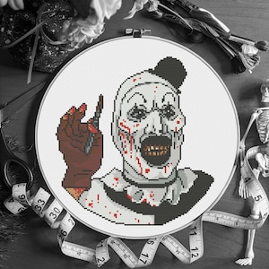 May include: A cross-stitch hoop featuring a pixelated image of a scary clown with blood splatter and a brown glove holding a knife. The clown has a black top hat and a wide, open mouth. The hoop is surrounded by measuring tape and embroidery floss.