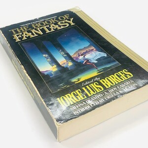 Może przedstawiać: Książka w twardej oprawie zatytułowana "THE BOOK OF FANTASY" z ilustracją na okładce przedstawiającą kobietę w białej szacie biegnącą w kierunku niebieskiego i złotego krajobrazu. Zredagował Jorge Luis Borges.
